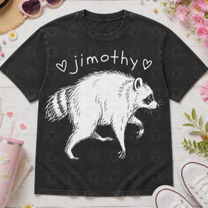 May include: A black short-sleeve t-shirt featuring a white line-art illustration of a raccoon in profile. Above the raccoon, the text "jimothy" is printed in a white handwritten font, flanked by two small white heart outlines on each side.