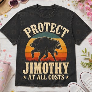 May include: A black short-sleeve t-shirt featuring a graphic of a raccoon centered over a sunset design. The text reads PROTECT JIMOTHY AT ALL COSTS in a bold, light-colored font. The shirt has a crew neckline and a standard, relaxed fit.