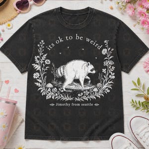 May include: A black short-sleeve t-shirt featuring a white graphic of a raccoon surrounded by a floral wreath with mushrooms. The text "its ok to be weird" is printed above the raccoon, and "Jimothy from seattle" is printed below the graphic.
