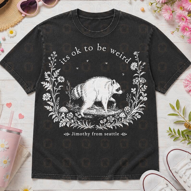 May include: A black short-sleeve t-shirt featuring a white graphic of a raccoon surrounded by a floral wreath with mushrooms. The text "its ok to be weird" is printed above the raccoon, and "Jimothy from seattle" is printed below the graphic.