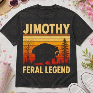 May include: A black short-sleeve t-shirt featuring a graphic print of a raccoon silhouette against a sunset background with trees. The text JIMOTHY is printed in yellow above the graphic, and the text FERAL LEGEND is printed in yellow below.