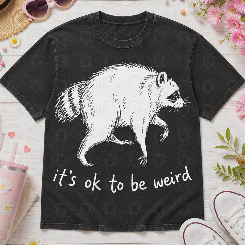 May include: A black short-sleeve t-shirt featuring a white line-art illustration of a raccoon in the center. Below the raccoon, the text reads "it's ok to be weird" in a simple white font. The shirt has a crew neckline and a relaxed fit.