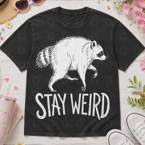 May include: A black short-sleeve t-shirt with a white graphic illustration of a walking raccoon in the center. Below the raccoon, the text STAY WEIRD is printed in a white, stylized, hand-drawn font. The shirt has a crew neckline and a faded finish.