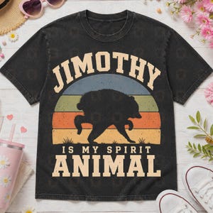 May include: A black short-sleeve t-shirt featuring a graphic of a black bear silhouette centered within a vintage-style sunset of blue, green, orange, and yellow stripes. The text reads JIMOTHY IS MY SPIRIT ANIMAL in a bold, light-colored font.