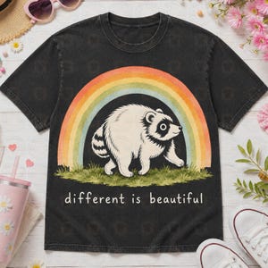 May include: A black short-sleeve t-shirt featuring a graphic of a white raccoon standing on green grass in front of a multi-colored rainbow arch. The text "different is beautiful" is printed in white letters below the raccoon illustration.