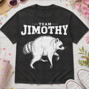 May include: A black short-sleeve t-shirt featuring a white graphic of a raccoon in profile. Above the raccoon, the text reads "TEAM JIMOTHY" in a bold, white, capitalized font. The shirt has a crew neckline and a slightly faded, vintage-style finish.