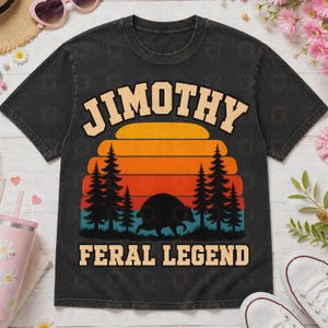 May include: A black short-sleeve t-shirt featuring a graphic of a sunset behind pine trees and a silhouette of a bear. The text JIMOTHY is printed in a tan arched font above the graphic, and FERAL LEGEND is printed in tan below the graphic.