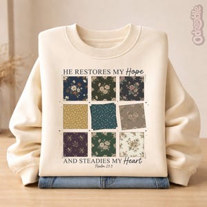 May include: Cream-colored sweatshirt with the text "HE RESTORES MY HOPE AND STEADIES MY HEART" and Psalm 23:3. The design features nine floral pattern squares in various colors. The sweatshirt is folded over a pair of blue jeans.