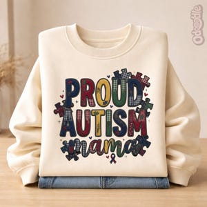 May include: Cream-coloured sweatshirt featuring the text "PROUD AUTISM mama" in colourful, patterned letters and puzzle piece accents. The sweatshirt is folded over a pair of blue jeans.
