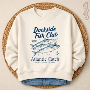 Puede incluir: Sudadera color crema con el texto "Dockside Fish Club" y "Atlantic Catch" en azul. El diseño presenta tres peces, un nudo marinero, un ancla y estrellas. La sudadera se muestra sobre una cesta tejida.