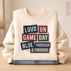 Könnte beinhalten: Ein cremefarbenes Sweatshirt mit dem Text "LOUD ON GAME DAY BLUE THROUGH & THROUGH" in marineblauen, roten und grauen Rechtecken. Das Sweatshirt ist über einer Jeans gefaltet.