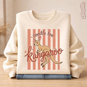 Puede incluir: Sudadera de color crema con una ilustración de un canguro de dibujos animados y las palabras "toodle loo kangaroo". El canguro es de color marrón claro, sobre un fondo de rayas verticales melocotón. La sudadera está doblada sobre unos vaqueros azules.