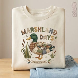 May include: Cream-coloured sweatshirt with a duck illustration. The duck has patchwork-style feathers and the text "MARSHLAND DAYS". The design includes cattails, dragonflies, and the words "FIELD & FEATHER" on a banner. The sweatshirt is folded on top of blue jeans.