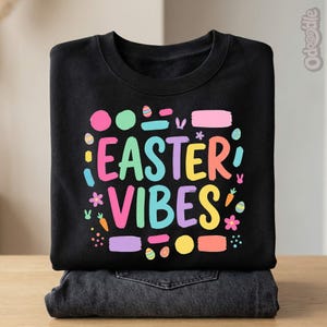 May include: Black sweatshirt featuring the text "EASTER VIBES" in a vibrant, multi-coloured design, with Easter-themed illustrations. The sweatshirt is folded on top of a pair of dark-wash jeans. Includes eggs, carrots, and flowers.