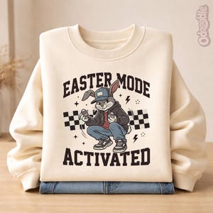 以下が含まれることがあります： 「EASTER MODE ACTIVATED」の文字と、帽子とジャケットを着てイースターエッグを持っているウサギのグラフィックが描かれた、クリーム色のスウェットシャツ。スウェットシャツは、青いジーンズの上に折りたたまれています。
