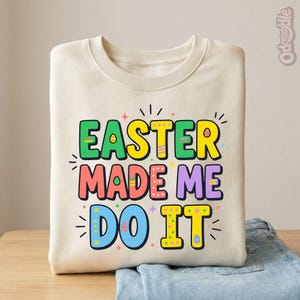 May include: A cream-colored sweatshirt with the text "EASTER MADE ME DO IT" in colorful, playful lettering. The design features green, yellow, pink, purple, and blue colors, with decorative accents. The sweatshirt is folded on top of a pair of blue jeans.