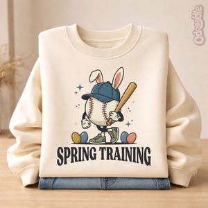 May include: Cream-colored sweatshirt with a baseball-themed graphic. The graphic features a baseball character wearing a baseball cap and bunny ears, holding a bat, with the text "SPRING TRAINING" below. Two blue and yellow Easter eggs are also present.