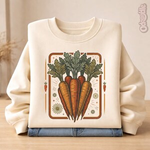 Puede incluir: Sudadera color crema con un gráfico de zanahorias. La ilustración presenta un grupo de zanahorias naranjas con hojas verdes, enmarcado en un rectángulo marrón con detalles florales. La sudadera está doblada sobre unos vaqueros azules.