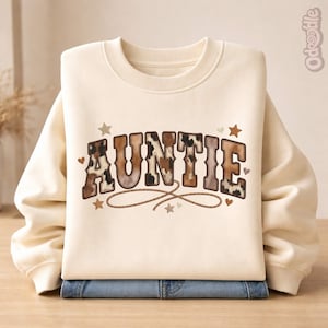 Puede incluir: Sudadera color crema con la palabra "AUNTIE" en marrón, con un diseño de piel de vaca. El diseño incluye estrellas, corazones y un detalle en forma de cuerda. La sudadera está doblada sobre unos vaqueros azules.
