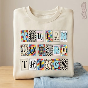May include: Cream-colored sweatshirt with the phrase "YOU CAN DO HARD THINGS" in colorful, block letter designs. The letters feature various patterns, including puzzle pieces, polka dots, and animal prints. The sweatshirt is folded on top of a pair of blue jeans.