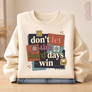 May include: Cream-colored sweatshirt with the text "don't let the hard days win" in a retro design. The graphic includes floral and heart accents. The sweatshirt is folded over a pair of blue jeans.