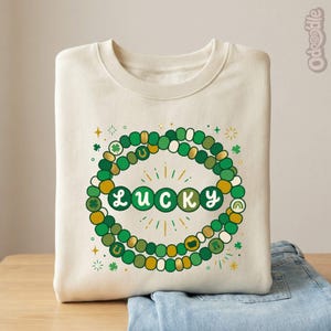 May include: Cream-colored sweatshirt with a St. Patrick's Day design. The design features a beaded circle in shades of green and gold, with the word "LUCKY" in green letters. The design also includes shamrocks, horseshoes, and a rainbow.
