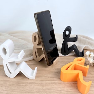 Könnte beinhalten: Eine Sammlung von vier stilisierten, menschenförmigen Handyhaltern in Weiß, Beige, Schwarz und Orange. Jeder Halter hat das Design einer sitzenden Figur mit einem ausgestreckten Arm, um ein Smartphone zu stützen.