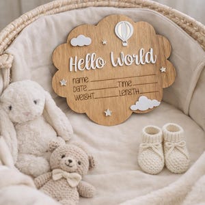 以下が含まれることがあります： 「Hello World」の文字と赤ちゃんの詳細を記入するスペースがある木製のサイン。雲、星、熱気球で装飾されています。うさぎのぬいぐるみ、テディベア、ニットのベビーブーツが入った柳のバスケットに入っています。