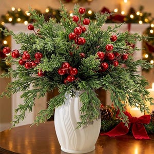 May include: A white ceramic vase with a vertical ribbed texture holds a dense arrangement of green pine branches and clusters of bright red berries. The foliage is full and textured, creating a rounded shape that fills the top of the vase.