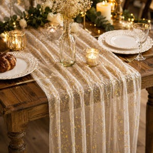 May include: A sheer white table runner featuring a scattered gold foil pattern draped over a wooden table. The runner is accented by a clear glass vase filled with dried flowers, gold-toned candle holders, and white plates on the table surface.