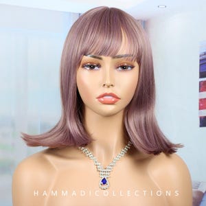 May include: A mannequin head with a mauve-colored wig featuring bangs and a shoulder-length cut. The mannequin wears a silver necklace with a blue teardrop-shaped pendant. The text "HAMMADICOLLECTIONS" is visible at the bottom.