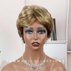May include: A mannequin head wearing a short, layered blonde wig, a sparkling rhinestone necklace, and matching earrings. The mannequin has dark eye makeup and glossy red lipstick. The text "HAMMARI COLLECTIONS" is visible at the bottom.