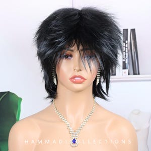 Peut inclure: Perruque noire à mèches courtes et ébouriffées. Elle est présentée sur une tête de mannequin, ornée d'un collier en strass étincelant et de boucles d'oreilles assorties. Le collier comporte un pendentif bleu en forme de goutte. Le texte "HAMMADI COLLECTIONS" est visible.