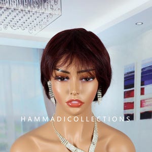 May include: A burgundy-colored wig with a short, layered style. The image also features a sparkling silver necklace and matching earrings. The text "HAMMADICOLLECTIONS" is visible at the bottom of the image.