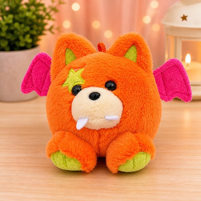 May include: An orange plush toy with pink wings, green accents, and a star. The toy has a bear-like face with black eyes, a white snout, and two fangs. A small, red loop is at the top of the head. The toy is sitting on a wooden surface.