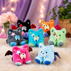 May include: A collection of plush bear toys in various colors, including black, gray, orange, green, pink, and blue. Each bear has bat wings, a star-shaped patch, and a small fang. The bears are sitting on a soft, white surface.