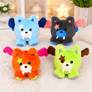 May include: Four plush toys in blue, gray, orange, and green, each with bat-like wings and a star accent. The toys have white fangs and are designed to resemble a bear or dog. The toys are sitting on a white, fluffy surface.
