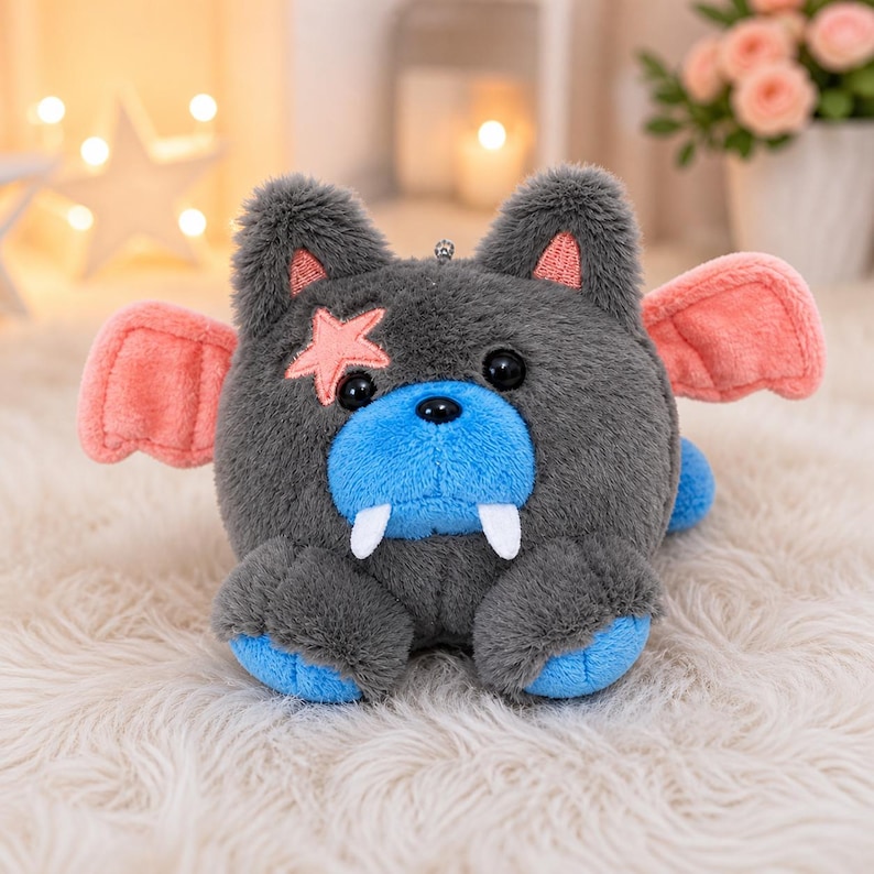 May include: A plush toy creature with gray fur, blue snout and paws, and pink accents on the ears, wings, and a star-shaped patch. The toy has large black eyes and two white fangs. It is sitting on a white, fluffy surface.