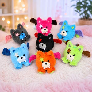 May include: A collection of plush toys in various colors, including pink, blue, orange, green, black, and gray. Each toy has a bear-like face with fangs, star accents, and bat wings. The toys are arranged on a soft, pink surface.