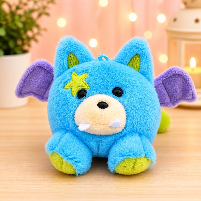 May include: A plush toy creature in shades of blue, purple, and green. The round-bodied toy has large ears, small wings, and a star-shaped patch. It features a light-colored snout, two small fangs, and black eyes. The toy is designed to sit upright.