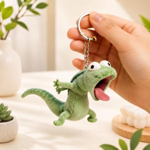 May include: A green plush lizard keyring with a wide open mouth and pink tongue. The lizard has large, googly eyes and is attached to a silver keyring. The keyring is held up by a hand.