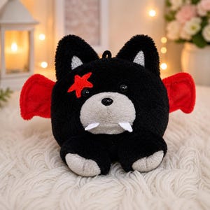 May include: A plush toy in the shape of a black bear with red wings, a red star, and white fangs. The bear has a gray snout and paws. The toy is sitting on a white, fluffy surface.
