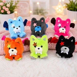 May include: Six colorful plush toys shaped like bears with bat wings. The bears are blue, gray, pink, orange, green, and black. Each bear has a star-shaped patch and white fangs. The toys are sitting on a pink blanket.