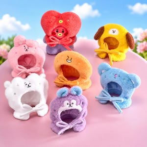 Puede incluir: Colección de siete sombreros de peluche de personajes en varios colores, incluyendo rojo, amarillo, rosa, blanco, naranja, azul y morado. Cada sombrero presenta un diseño único con una cara y un lazo. Los sombreros se muestran sobre una superficie rosa con un fondo de cielo.