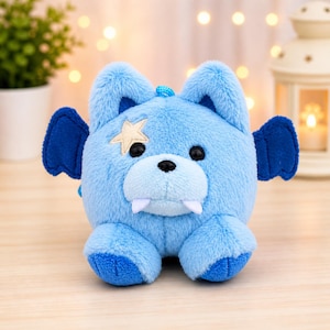 May include: A light blue plush toy with a bear-like face, bat wings, and a star-shaped patch. The toy has black eyes, white fangs, and blue accents. It is a soft, cuddly creature.