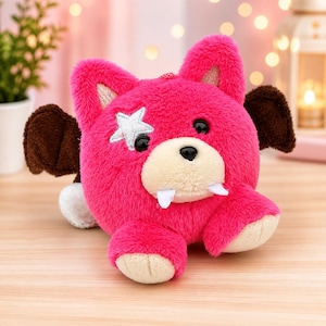 May include: A plush toy in the shape of a pink bear with brown bat wings. The bear has a white star on its face, black eyes, and two white fangs. The toy is designed to look like a cute, cuddly creature.