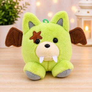 May include: A lime green plush toy with a cartoonish design. It features brown bat wings, a brown star on one eye, and two white fangs. The toy has black eyes, gray accents, and a green loop for hanging.