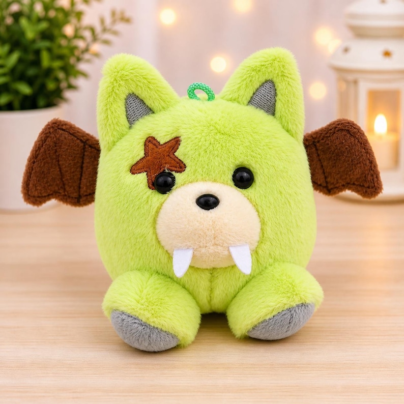 May include: A lime green plush toy with a cartoonish design. It features brown bat wings, a brown star on one eye, and two white fangs. The toy has black eyes, gray accents, and a green loop for hanging.