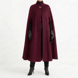 Peut inclure: Une cape bordeaux avec un col montant et un fermoir doré. La longue cape fluide est portée sur une tenue noire, comprenant des gants et des bottes. La cape est un vêtement d'extérieur élégant.