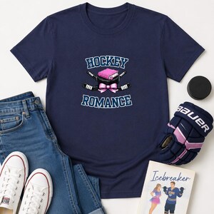 May include: A navy blue t-shirt featuring a graphic of two crossed hockey sticks, a pink book, and a pink bow, with the text HOCKEY ROMANCE printed in white and blue. The shirt is displayed with blue jeans, white sneakers, a hockey glove, and a book.